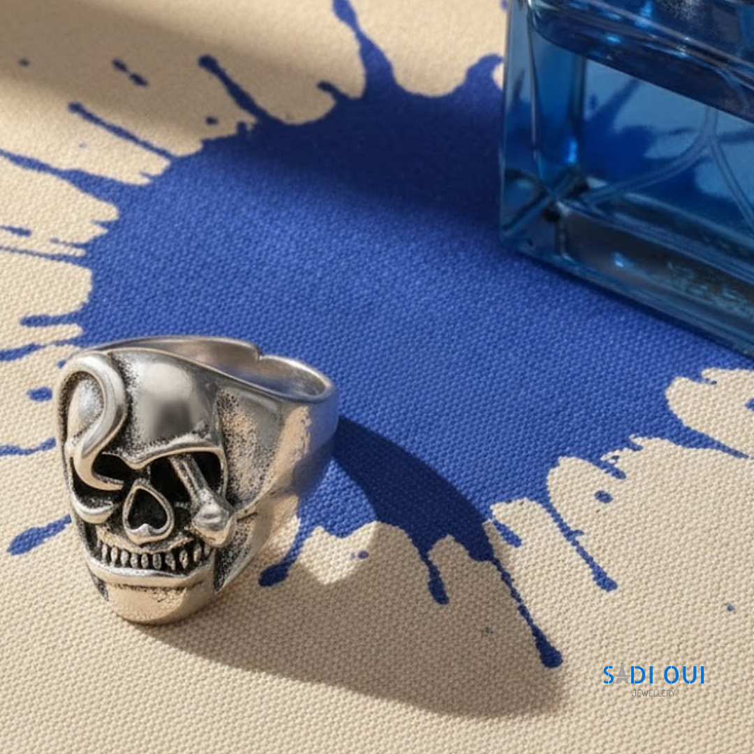Anel Caveira Base em aço inoxidável prateado em fundo bege e azul com mancha de líquido azul

